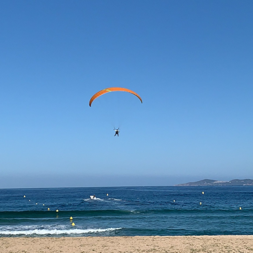  Individu s'envolant dans les airs à bord d'un parapente dans plage de Liscia en Corse. Plage avec des vagues, du sable. Montagnes avec végétation au loin. 