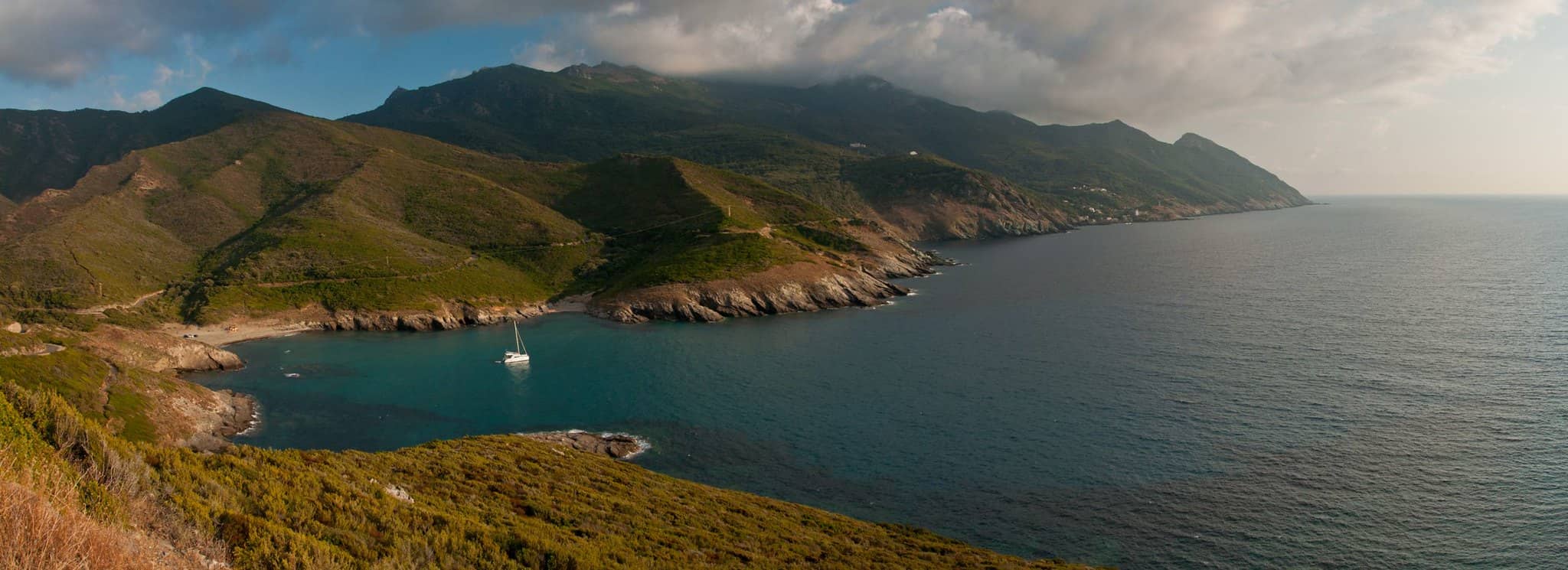 Paysage du Cap Corse. Bateau naviguant dans la mer bleu turquoise entouré de montagnes et de végétation.