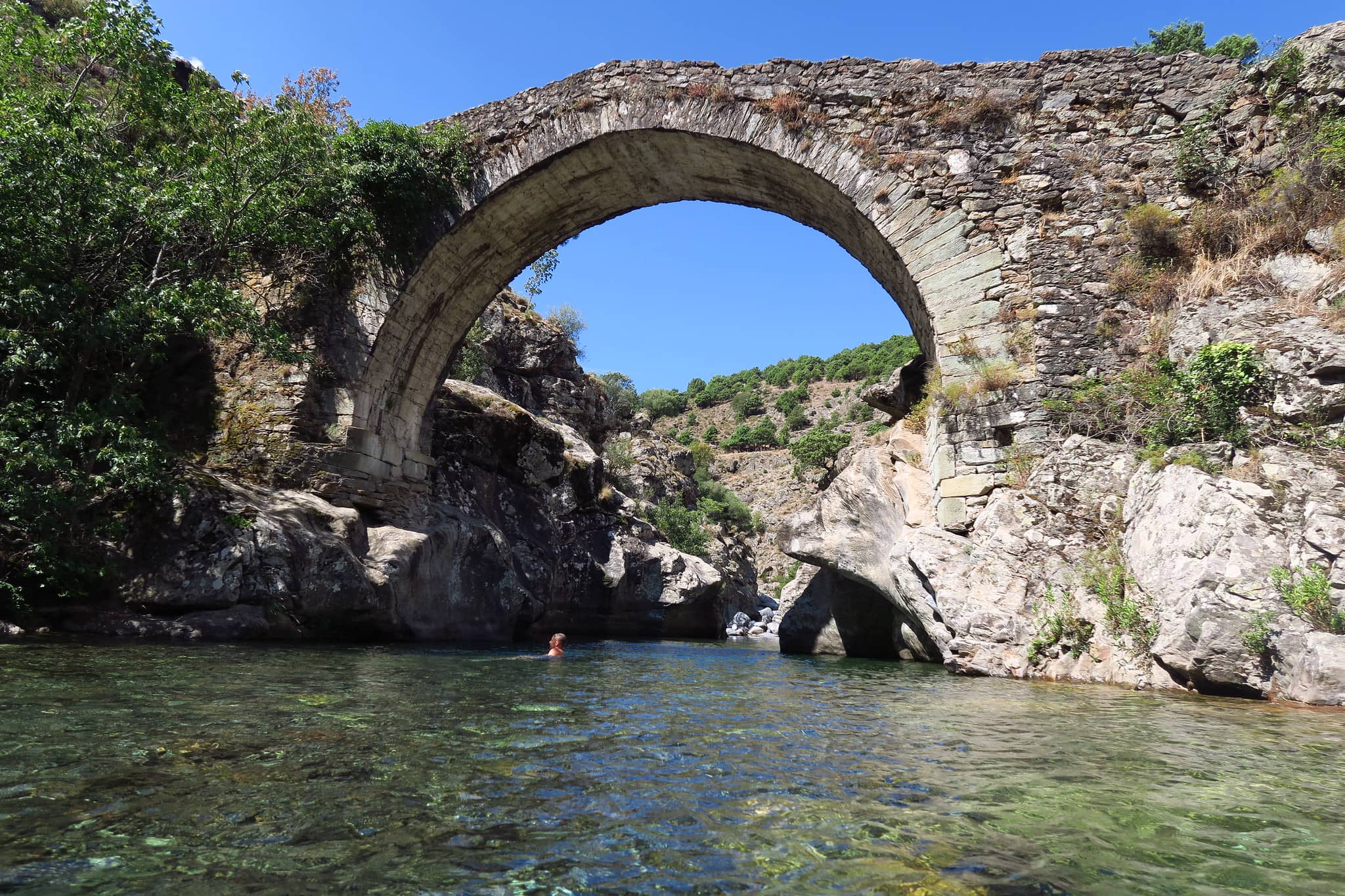 Photo du pont génois et les roches encadrant la piscine naturelle émeraude. Arche en pierres sous forme de voûte. Présence d'une silhouette humaine dans la piscine naturelle.