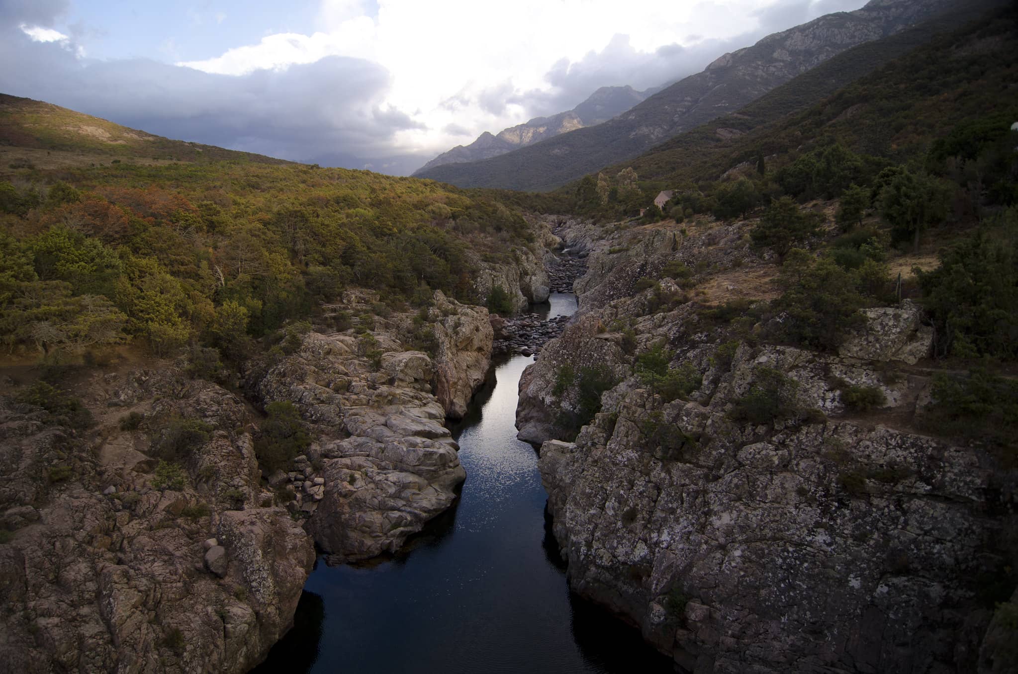 Vue panoramique de la vallée du Fango. Présence de blocs de roches encadrant le fleuve, végétation, montagnes au loin. 