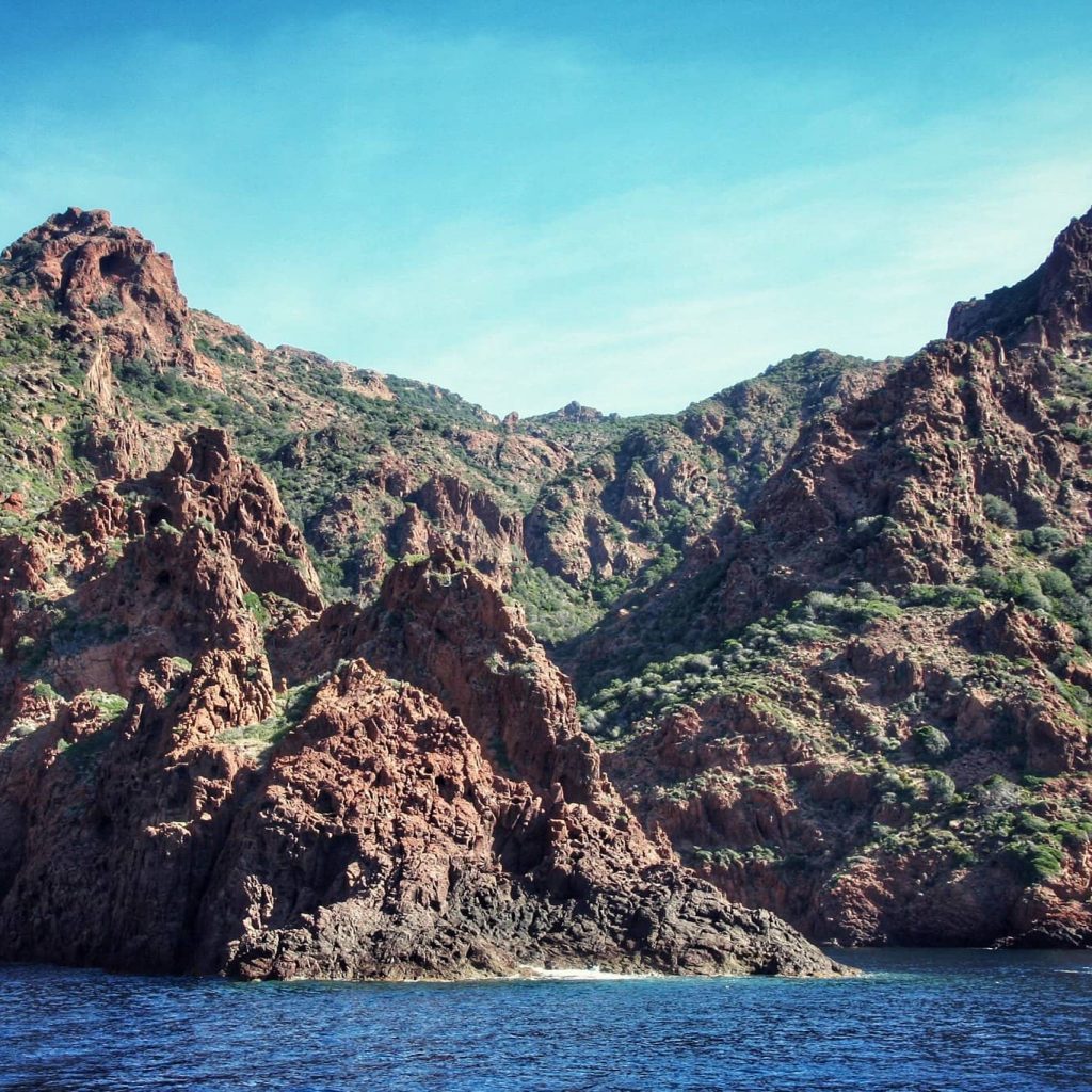 Paysage de la réserve naturelle de Scandola. Présence de falaises de porphyre rouge avec végétation verte sur la mer bleue où navigue un bateau. 